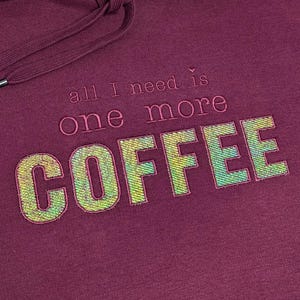 Könnte beinhalten: Ein weinroter Kapuzenpullover mit Kordelzug und dem gestickten Text "all I need is one more COFFEE." Das Wort "COFFEE" ist schimmernd grün und gelb.