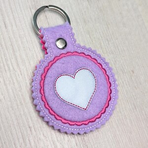 Könnte beinhalten: Lilafarbener Schlüsselanhänger aus Filz mit einem rosa gewellten Rand und einem weißen Filzherz in der Mitte. Der Schlüsselanhänger hat einen silbernen Metallring.