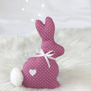 Può includere: Peluche coniglio rosa a pois bianchi con un fiocco bianco e un cuore bianco sul ventre. Il coniglio è seduto su una superficie bianca e soffice.