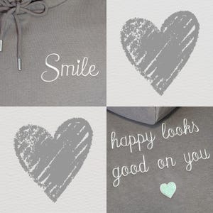 Op de afbeelding: Een grijze hoodie met witte trekkoorden en het woord "Smile" in cursief. Twee grijze harten met een textuurontwerp op een witte achtergrond. De woorden "happy looks good on you" zijn ook zichtbaar.