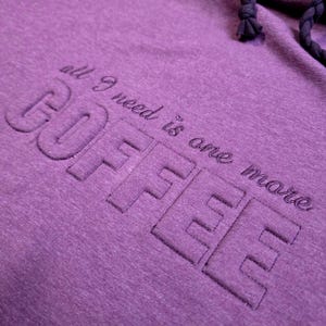 Könnte beinhalten: Nahaufnahme eines lila Hoodies mit dem gestickten Text "COFFEE" in großen Blockbuchstaben. Über dem Wort "COFFEE" steht der gestickte Text "all I need is one more". Der Hoodie hat einen Kordelzug.