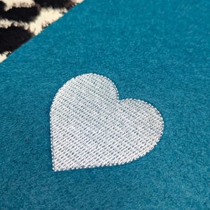 Könnte beinhalten: Ein türkisfarbener Stoff mit einem silbernen, gestickten Herz. Das Herz ist mit einem strukturierten, gewebten Muster gefüllt. Der Hintergrund ist einfarbig türkis, mit einem Teil eines schwarz-weiß gemusterten Stoffes in der oberen linken Ecke.