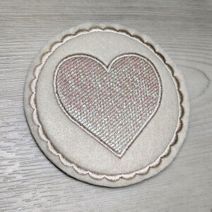 May include: A round, off-white coaster with a heart design. The heart is filled with a shimmering, textured fabric and outlined with a light brown thread. The coaster has a scalloped edge.