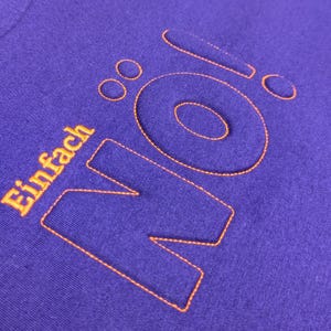Könnte beinhalten: Lilafarbenes T-Shirt mit dem Wort "NO" in orangefarbener Stickerei. Das Wort "einfach" ist ebenfalls in orangefarbener Stickerei unter dem Wort "NO" gestickt.