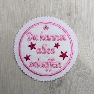 Könnte beinhalten: Ein weißer Filzkreis mit gewelltem Rand, mit einem rosa gestickten Design. Das Design umfasst einen rosa kreisförmigen Rand, den deutschen Text "Du kannst alles schaffen" und vier rosa Sterne.