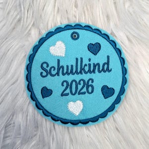 Può includere: Un ornamento rotondo in feltro azzurro con un bordo smerlato blu scuro. Il testo "Schulkind 2026" è ricamato in blu scuro. Cuori bianchi e blu scuro sono sparsi intorno al testo.