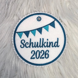 Könnte beinhalten: Ein weißer, runder Aufnäher mit einem gewellten Rand und einem türkisblauen Design. Der Aufnäher zeigt den Text "Schulkind 2026" in türkisfarbener Schrift, mit einem Banner aus Wimpeln.