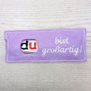 May include: A purple felt gift bag with a white stitched message that says "du bist großartig!" which translates to "you are great!" in English. The bag has a cut-out window to reveal a chocolate bar.