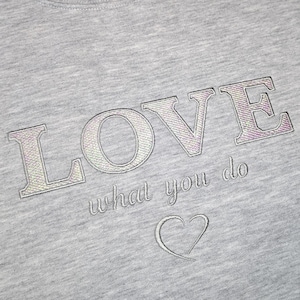 May include: A close-up shot of a grey sweatshirt featuring the embroidered words "LOVE what you do" in iridescent thread. A small heart is embroidered below the text. The fabric has a soft, textured appearance.