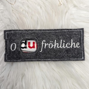 May include: A grey felt Christmas ornament with a white stitched border. The ornament has a small window with a red and white candy bar inside. The text "O du fröhliche" is stitched in white on the ornament.