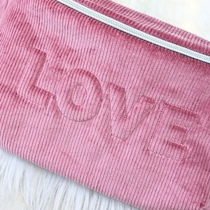 Puede incluir: Una bolsa de cremallera de pana rosa con la palabra "LOVE" bordada en hilo blanco.