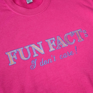 Könnte beinhalten: Ein leuchtend pinkfarbenes Sweatshirt mit dem gestickten Text "FUN FACT: I don't care!" in blauer und goldener Schrift. Das Sweatshirt hat einen Rundhalsausschnitt und lange Ärmel.