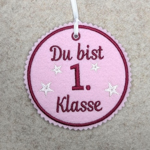 Könnte beinhalten: Runde Dekoration aus rosa Filz mit dem Text "Du bist 1. Klasse" in burgunderfarbener Schrift. Die Dekoration hat einen gewellten Rand, weiße Sternakzente und ein weißes Band zum Aufhängen.