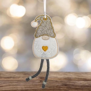 Könnte beinhalten: Ein Weihnachts-Wichtel-Ornament mit glitzerndem silbernen Hut und weißem Bart. Der Wichtel hat ein gelbes Herz auf der Brust und graue, herabhängende Beine. Ein weißes Band ist zum Aufhängen befestigt. Das Ornament ist für die Weihnachtsdekoration gedacht.