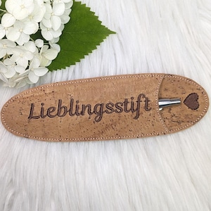Puede incluir: Un portaplumas de corcho marrón con el texto "Lieblingsstift" bordado. Un bolígrafo plateado está dentro del portaplumas.