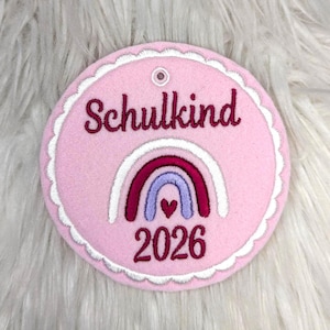 Könnte beinhalten: Runder, rosa Filzschmuck mit dem Wort "Schulkind" in bordeauxfarbener Schrift. Ein Regenbogen-Design mit Herz und die Jahreszahl "2026" in Bordeaux. Mit einer gewellten Kante und einem kleinen Loch.