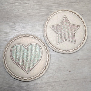 Könnte beinhalten: Zwei beige Stoffuntersetzer mit gestickten Designs. Einer zeigt ein Herz, der andere einen Stern. Beide haben einen gewellten Rand und eine strukturierte, irisierende Mitte.