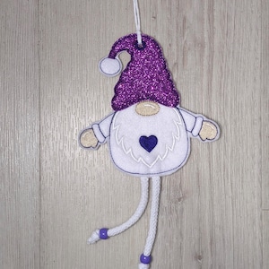 May include: A hanging gnome ornament crafted from white felt with a purple glitter hat, a beige nose, and beige hands. The gnome features a dark purple heart embroidered on its white beard and two dangling white cord legs with purple beads.