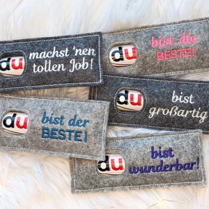 Puede incluir: Cinco bolsas de regalo de fieltro gris con un logotipo rojo, blanco y azul que dice "du" y mensajes bordados en diferentes colores. Los mensajes son "machst 'nen tollen Job!" en blanco, "bist die BESTE!" en rosa, "bist der BESTE!" en azul, "bist großartig!" en blanco y "bist wunderbar!" en morado.