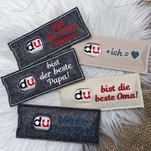 Könnte beinhalten: Fünf Filzgeschenktüten mit bestickten Botschaften. Die Taschen sind grau und beige und zeigen das Wort "du" in einem Kreis mit einem rot-blauen Design. Die Botschaften auf den Taschen lauten: "du bist die beste Mama!"  "du bist der beste Papa!" "du + ich = "  "du bist die beste Oma!"  "du bist der beste Opa!"