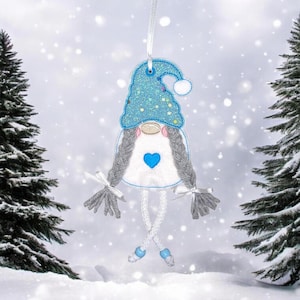 Puede incluir: Adorno de gnomo caprichoso con un sombrero azul brillante y un cuerpo blanco adornado con un corazón azul. El gnomo tiene trenzas grises y piernas colgantes, perfecto para la decoración navideña. Cuelga de una cinta blanca.