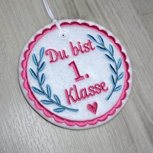 Peut inclure: Décoration ronde en feutre blanc, bordée d'un feston rose et d'une ficelle blanche. Le texte "Du bist 1. Klasse" est brodé en rose, avec des accents de feuilles bleues et un cœur rose.