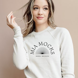 以下が含まれることがあります： 「MIA MOCKS MODEL MOCKUPS」と黒で書かれたグラフィックデザインが施された白い長袖スウェットシャツ。太陽の光が放射状に広がっているデザインです。