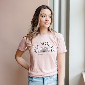 Op de afbeelding: Een vrouw draagt een roze T-shirt met de tekst "MIA MOCKS MODEL MOCKUPS" erop gedrukt. Ze staat voor een raam.