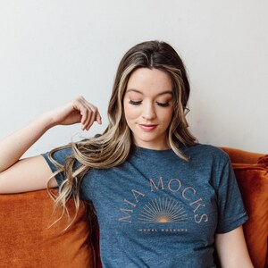 Op de afbeelding: Een vrouw draagt een donkerblauw T-shirt met de tekst "MIA MOCKS MODEL MOCKUPS" in roze letters. Ze zit op een oranje bank.