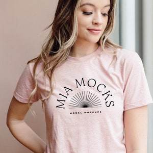 Op de afbeelding: Een roze T-shirt met een zwart grafisch ontwerp met de tekst "MIA MOCKS MODEL MOCKUPS" met een zonnestralenontwerp.