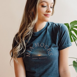 Op de afbeelding: Een vrouw draagt een donkerblauw t-shirt met een roze en gouden zon-graphic en de tekst "Mia Mocks Model Mockups".