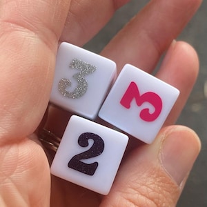 May include: Three white dice with different colored numbers. One die has a silver glitter number 3, another has a pink number 3, and the last die has a black number 2. The dice are held in a hand.