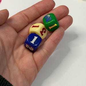 May include: Three colorful dice are held in a hand. One die is blue with white numbers, another is cream-colored with red numbers, and the third is green with yellow numbers. The dice are used for games.