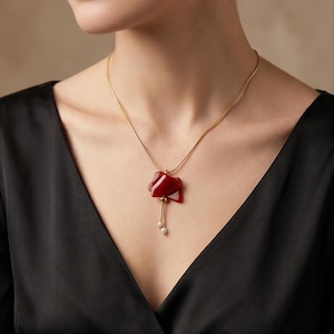 Peut inclure: Un collier doré avec un pendentif rouge abstrait et deux petites perles. Le collier est porté sur un haut noir. La forme du pendentif est le point central, avec les perles ajoutant une touche délicate.