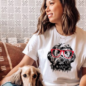 Puede incluir: Camiseta blanca con una ilustraci&oacute;n en blanco y negro de un perro con gafas rojas. La cara del perro es el punto central, con detalles de pelaje y las gafas que a&ntilde;aden un toque de color. La camiseta la lleva una persona.
