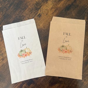 May include: Two paper favor bags, one white and one brown, with a fall-themed design. The bags feature the text "FALL in Love" above a watercolor image of pumpkins and flowers. The text "Jessica's Bridal Shower November 12th, 2025" is also printed.