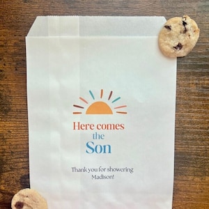 以下が含まれることがあります： 「Here comes the Son」の文字とオレンジ、ティール、赤色の太陽のグラフィックが入った白い紙袋。バッグには「Thank you for showering Madison!」の文字も含まれています。チョコレートチップクッキーが2つ、バッグの隣にあります。