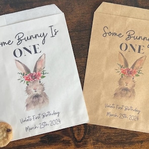 以下が含まれることがあります： 水彩画のウサギのデザインと「Some Bunny Is ONE」の文字が入った、白と茶色の2つの紙袋。袋はバイオレットの1歳の誕生日、2024年3月25日用です。チョコレートチップクッキーが各袋の近くにあります。