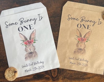 May include: Two paper treat bags, one white and one brown, with a watercolor bunny design and the text "Some Bunny Is ONE." The bags are for Violet's First Birthday, March 25th, 2024. A chocolate chip cookie rests near each bag.