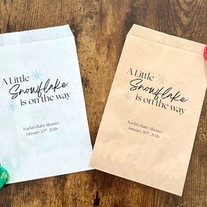 May include: Two paper treat bags with the text "A Little Snowflake is on the way" and baby shower details. One bag is white, the other is tan. Decorative candies are placed near the bags.