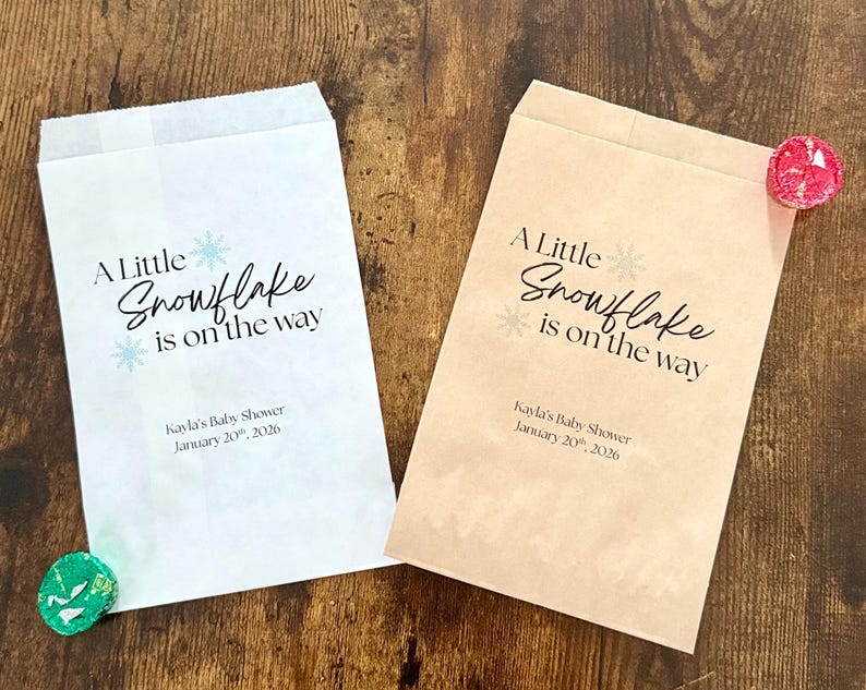 May include: Two paper treat bags with the text "A Little Snowflake is on the way" and baby shower details. One bag is white, the other is tan. Decorative candies are placed near the bags.