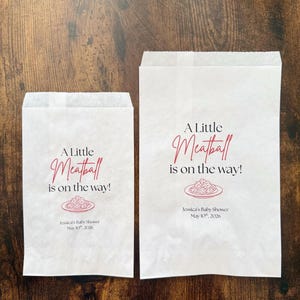 May include: Two white paper bags with the text "A Little Meatball is on the way!" in a script font. The bags also feature a plate of meatballs graphic and the text "Jessica's Baby Shower May 10th, 2026."