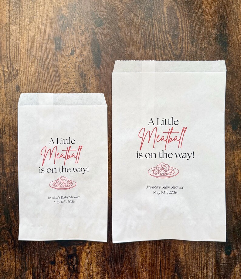 May include: Two white paper bags with the text "A Little Meatball is on the way!" in a script font. The bags also feature a plate of meatballs graphic and the text "Jessica's Baby Shower May 10th, 2026."
