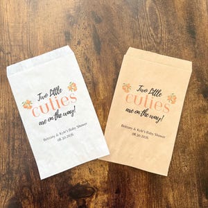 May include: Two paper treat bags, one white and one tan, with the text "Two Little Cuties are on the way!" in peach and dark gray. The bags are for Brittany & Kyle's Baby Shower, 08.30.2026.