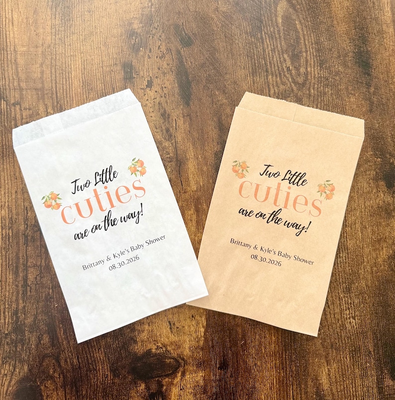 May include: Two paper treat bags, one white and one tan, with the text "Two Little Cuties are on the way!" in peach and dark gray. The bags are for Brittany & Kyle's Baby Shower, 08.30.2026.