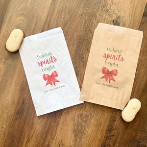 May include: Two rectangular paper bags, one white and one brown, each printed with the green and red text 'baking spirits bright' above a red bow illustration and the black text 'Love, The Smith Family', accompanied by two oval biscuits.