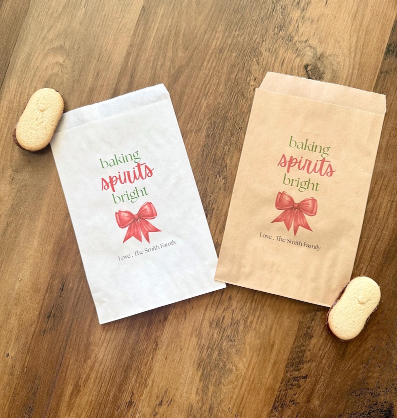 May include: Two rectangular paper bags, one white and one brown, each printed with the green and red text 'baking spirits bright' above a red bow illustration and the black text 'Love, The Smith Family', accompanied by two oval biscuits.