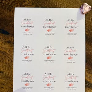 May include: A sheet of printed labels for a baby shower, featuring the text "A Little Sweetheart is on the way" in pink script. The labels also include the baby shower date and name.