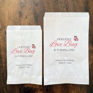 May include: Two white paper treat bags with the text "OUR LITTLE Love Bug IS TURNING ONE!" in pink script, a pink butterfly graphic, and the text "Jessica's First Birthday April 22nd, 2026".