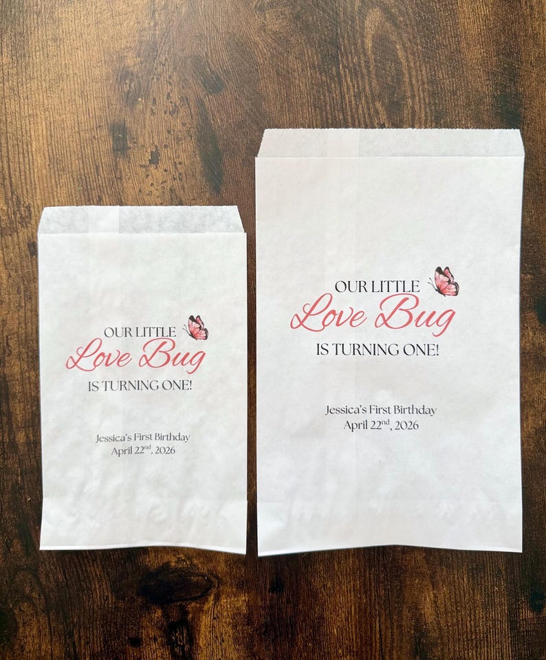 May include: Two white paper treat bags with the text "OUR LITTLE Love Bug IS TURNING ONE!" in pink script, a pink butterfly graphic, and the text "Jessica's First Birthday April 22nd, 2026".